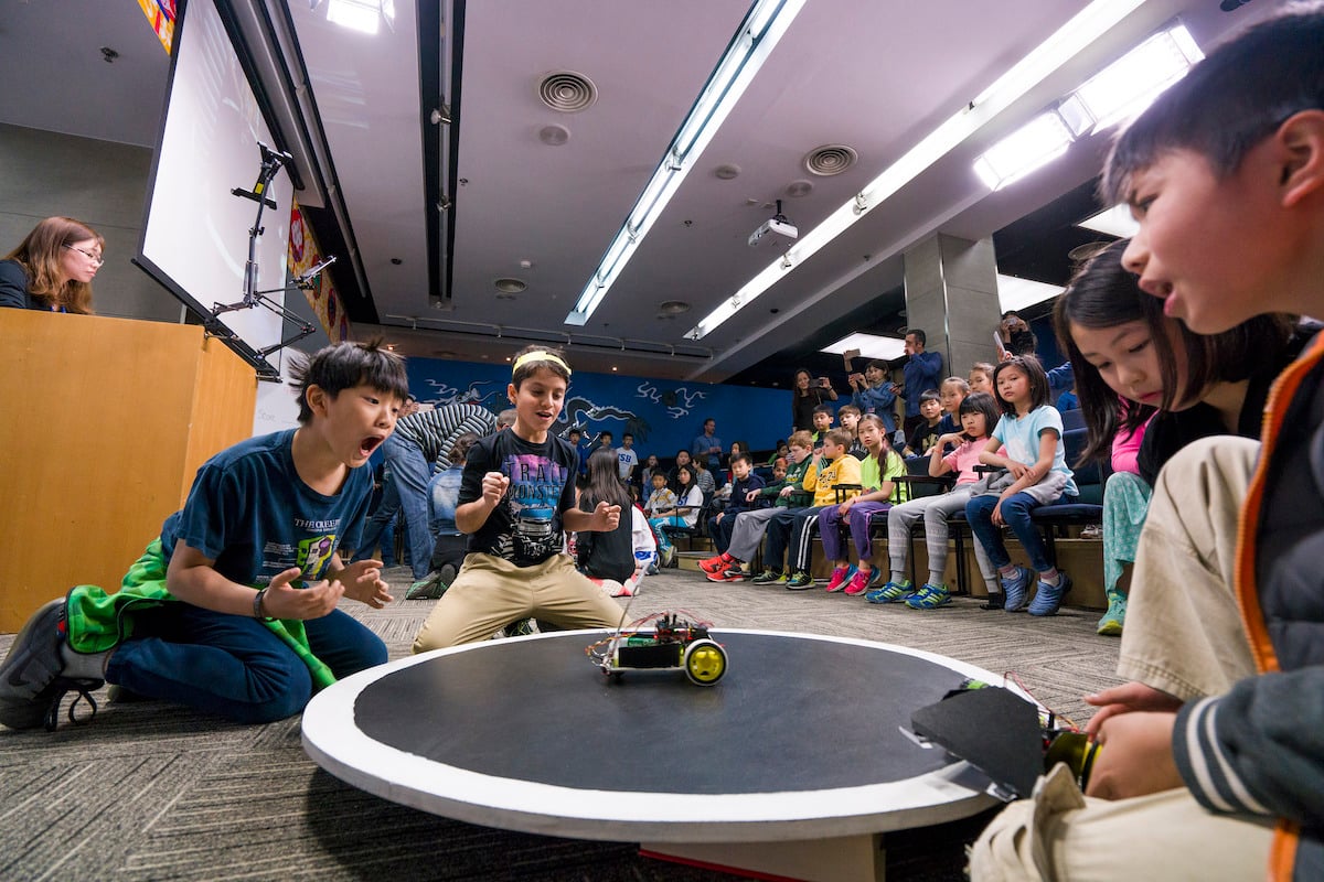 Students work excitedly with a robot game while others watch on in a theater